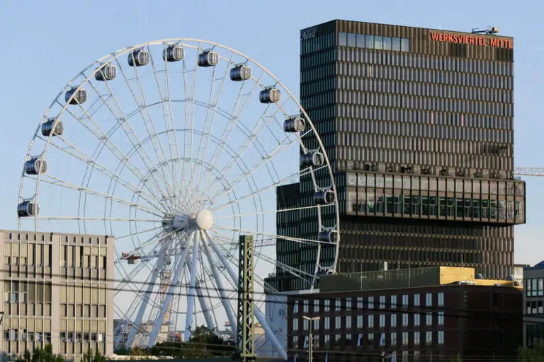 Das Umadum-Riesenrad im Münchner Werksviertel Mitte neben modernen Bürogebäuden und Baukränen, im Vordergrund sind Bahngleise sichtbar.