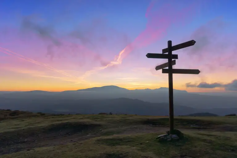 Dieses Bild zeigt eine ruhige Landschaft bei Sonnenuntergang oder Sonnenaufgang mit einem hölzernen Wegweiser, der mehrere Richtungen anzeigt. Der Wegweiser steht auf einer grasbewachsenen Anhöhe, im Hintergrund sind mehrere Bergketten zu sehen. Der Himmel erstrahlt in lebendigen Farben wie Rosa, Orange und Blau. Die Szene vermittelt ein Gefühl von Entscheidung, Orientierung oder einem Wendepunkt.
