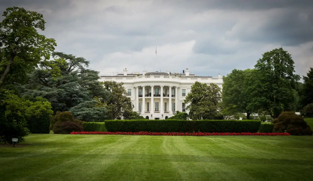 Das Weiße Haus in Washington, D.C., aufgenommen von der Gartenseite an einem bewölkten Tag. Der gepflegte Rasen und die symmetrische Anordnung von Bäumen und Blumen rahmen das ikonische Gebäude ein.