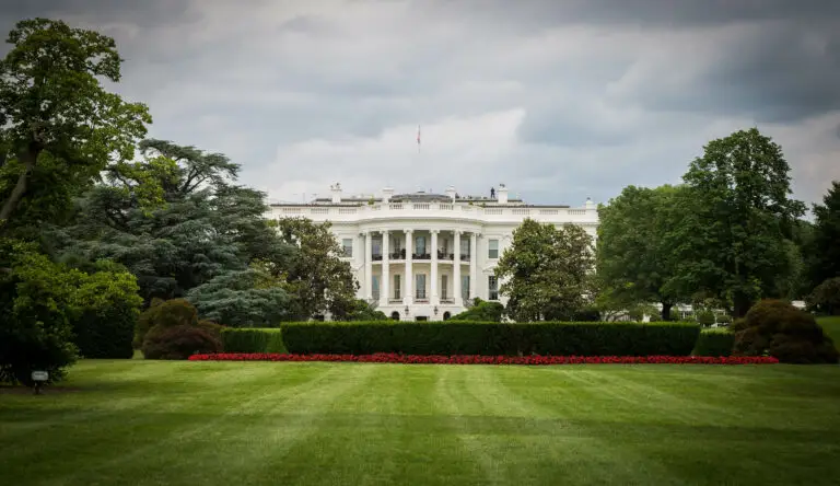 Das Weiße Haus in Washington, D.C., aufgenommen von der Gartenseite an einem bewölkten Tag. Der gepflegte Rasen und die symmetrische Anordnung von Bäumen und Blumen rahmen das ikonische Gebäude ein.