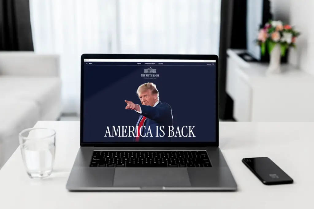 Ein Laptop steht auf einem weißen Tisch mit einem Glas Wasser und einem Smartphone daneben. Auf dem Bildschirm ist die Webseite des Weißen Hauses unter der zweiten Trump-Administration zu sehen, mit einem Bild von Donald Trump, der nach vorne zeigt, und dem Slogan "AMERICA IS BACK". Im Hintergrund ist ein helles, modern eingerichtetes Wohnzimmer zu erkennen.