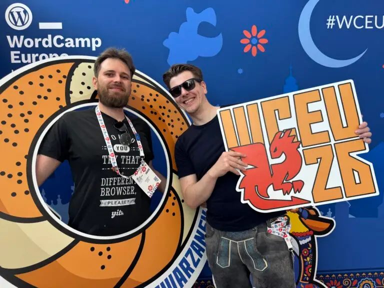 Zwei Männer posieren vor einer WordCamp-Europe-Fotowand; einer hält ein „WCEU 26“-Schild