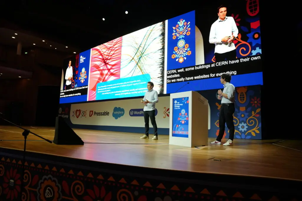 Zwei Sprecher auf einer WordCamp-Europe-Bühne vor einer großen Präsentationsleinwand mit CERN-Folien
